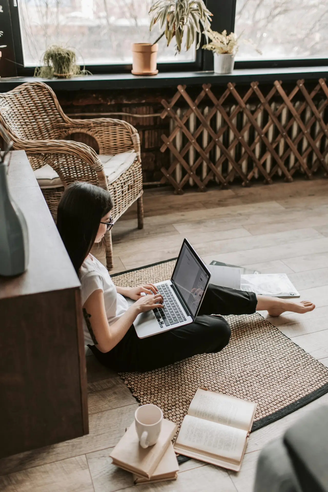 A young woman in casual attire working from home using a laptop while sitting on the floor with a coffee mug nearby.